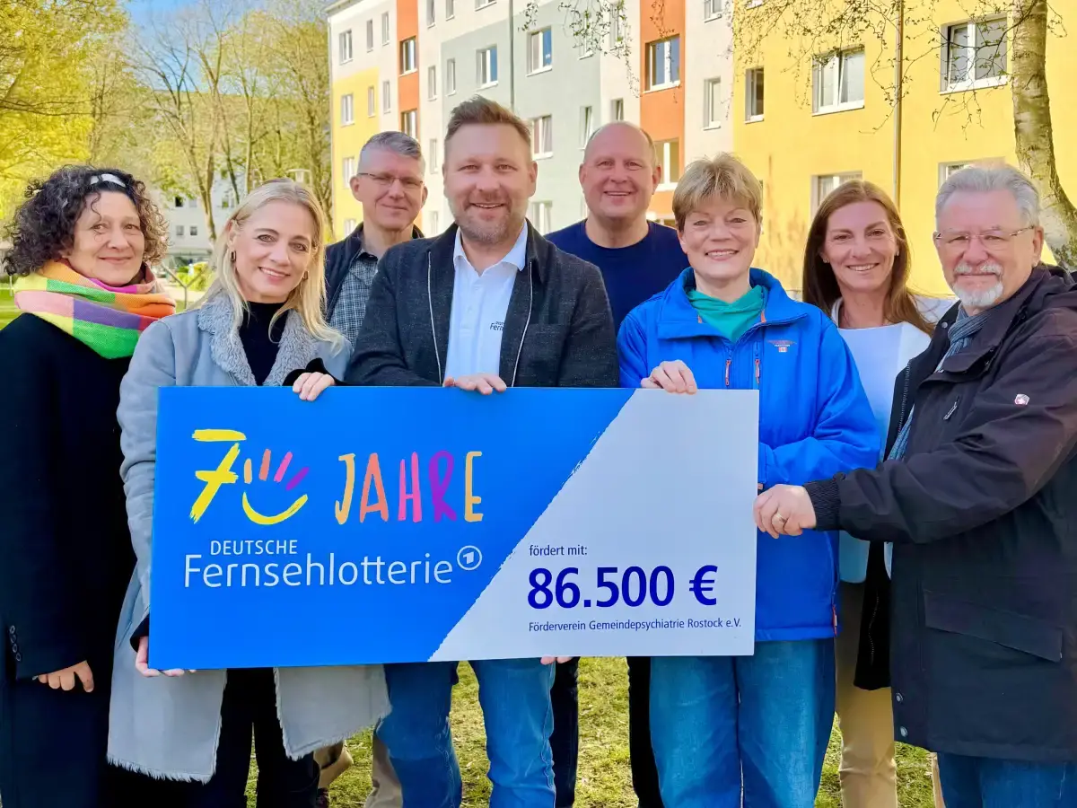 Acht Menschen stehen vor einem Wohnblock in der Rostocker S&uuml;dstadt und halten einen F&ouml;rderscheck der Deutschen Fernsehlotterie.